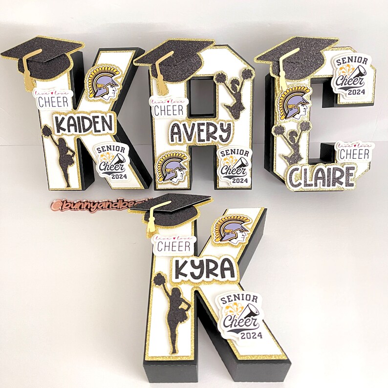 May include: Four black and white letter blocks with gold accents spelling out the word "KAKE" with each letter decorated with a cheerleader pom-pom, a graduation cap, a mascot helmet, and a "Senior Cheer 2024" sticker.