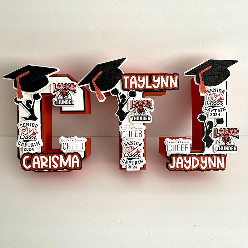 May include: A red, white and black foam board letter 'L' with the names Carisma, Taylynn and Jaydynn. The letter is decorated with graduation caps, cheerleading pom-poms and Lamar Thunder cheerleading logos.