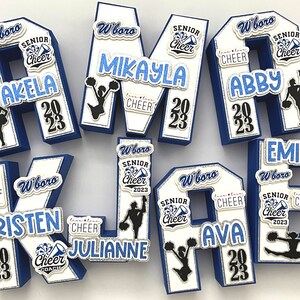 May include: A set of six blue and white foam letters spelling out the word "KELSEY" decorated with cheerleading-themed embellishments. Each letter has a white background with blue trim and features a cheerleader silhouette, the words "Senior Cheer", and the year "2023". The letters are decorated with glitter and other embellishments.