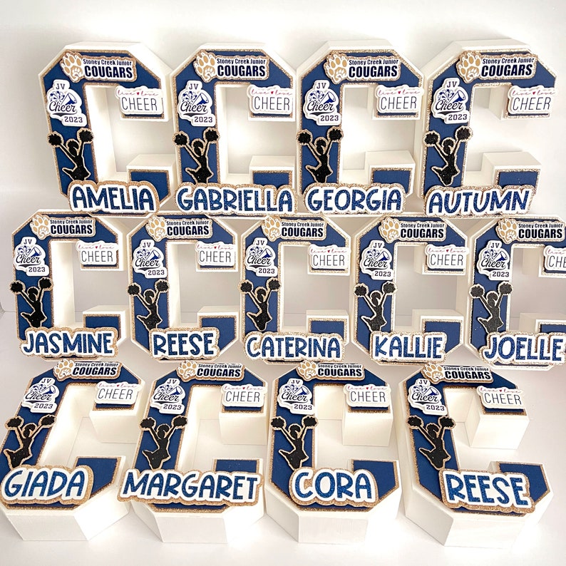 May include: A set of 10 white foam cheerleading numbers decorated with gold glitter and blue paint. Each number features a cheerleader silhouette and the text "Stoney Creek Junior Cougars JV Cheer 2023". The numbers are arranged in a row and spell out the names Amelia, Gabriella, Georgia, Autumn, Jasmine, Reese, Caterina, Kallie, Joelle, Giada, Margaret, Cora, and Reese.