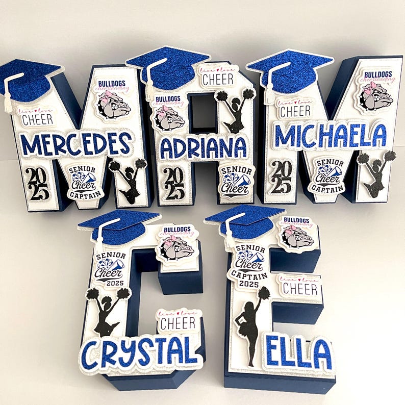 May include: Blue and white letter blocks spelling out the names "Mercedes", "Adriana", "Michaela", "Crystal", and "Ella". Each block features a graduation cap, a cheerleader silhouette, and a bulldog mascot. The blocks also include the text "Senior Cheer Captain 2025" and "Bulldogs Cheerleading".