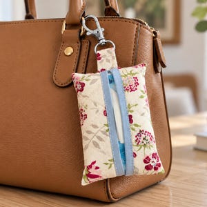 May include: A quilted fabric pouch with a floral pattern in shades of red, pink, and green, featuring a light blue center strip and a silver metal lobster clasp, hanging from the handle of a tan leather handbag.