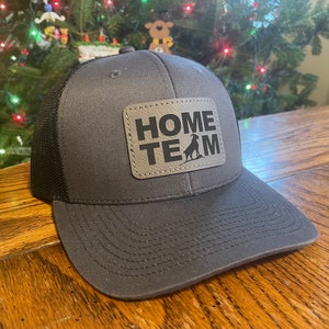 Peut inclure: Un chapeau de camionneur gris et noir avec un patch en cuir marron qui dit "HOME TEAM" avec une silhouette de chèvre.