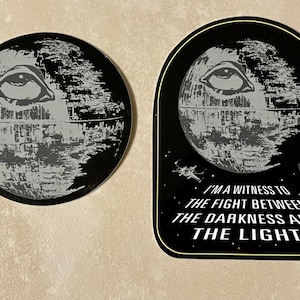 May include: Two black and white stickers featuring a Death Star with an eye in the center. The larger sticker has the text "I'm a witness to the fight between the darkness and the light".