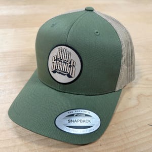 Può includere: Un cappello da camionista verde e beige con una toppa marrone che dice "Billy Strings" in un cerchio. Il cappello è uno snapback del marchio Yupoong.