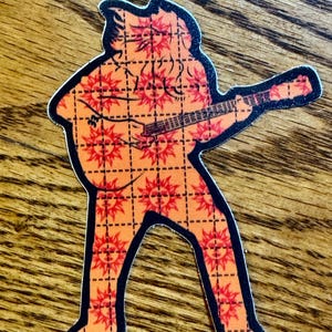 Peut inclure: Un autocollant découpé représentant une silhouette jouant de la guitare. La silhouette est orange avec un motif de soleil rouge répété et un contour noir. L'autocollant est sur une surface en bois.