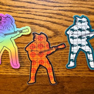 Peut inclure: Trois autocollants colorés représentant un musicien jouant de la guitare. Les silhouettes des musiciens sont remplies de motifs et de couleurs variés, dont un dégradé arc-en-ciel, un motif floral orange et un motif de véhicules verts.