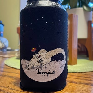 May include: Black can cooler with a graphic of a skull, a flower, and the text "bmfs" in a stylized font. The design is set against a starry night sky. The can cooler is designed to keep beverages cold.