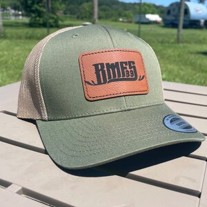 May include: A green and tan trucker hat with a brown leather patch that says "BMFS 1939".