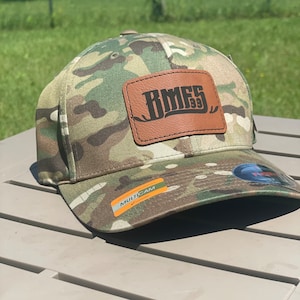 May include: Camouflage baseball cap with a brown leather patch displaying the text "RMF5 93". The cap has a curved peak and a sticker that says "MULTICAM".