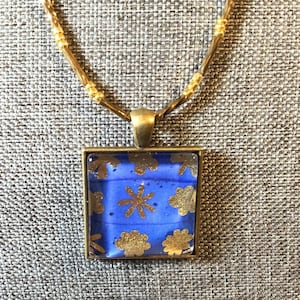 May include: A square pendant necklace with a blue background and gold floral designs. The pendant is framed in a gold-colored metal and hangs from a gold-toned chain. The necklace is displayed on a gray textured surface.