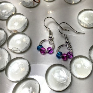 Puede incluir: Pendientes de aro plateados con cuentas azules y moradas. Los pendientes tienen un diseño sencillo con un pequeño aro plateado y un gancho para la oreja. Las cuentas son una mezcla de azul y morado.
