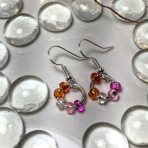 Puede incluir: Pendientes de aro plateados adornados con pequeñas cuentas de colores naranja, rosa y transparentes. Los pendientes se exhiben sobre una superficie blanca rodeada de piedras de vidrio transparentes y redondeadas.