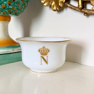 May include: A white porcelain bowl featuring a gold-colored crown emblem positioned above the letter N. The bowl has a smooth, curved silhouette with a delicate gold rim along the top edge and a matching gold band around the base.