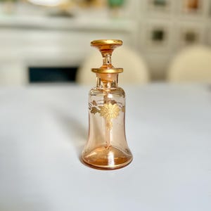 May include: A peach-colored glass perfume bottle with a gold-colored stopper and floral design. The bottle has a flared base and a slender neck. The floral design is gold and is located on the front of the bottle.