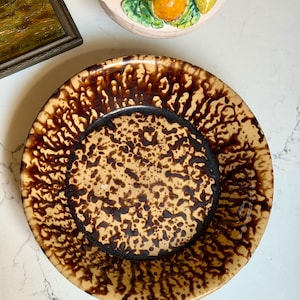 May include: A brown and tan ceramic plate with a speckled design. The plate has a dark brown rim and center, with a lighter tan background. The plate is likely vintage and suitable for serving food.