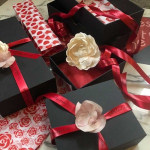 May include: Four black gift boxes wrapped in red ribbon with the words "Happy Birthday" and "Valentine's" printed on the ribbon. The boxes are filled with red and white tissue paper and decorated with white and pink flowers.