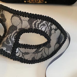 May include: A black lace masquerade mask with a black ribbon tie. The mask has a delicate floral pattern and a black satin trim.