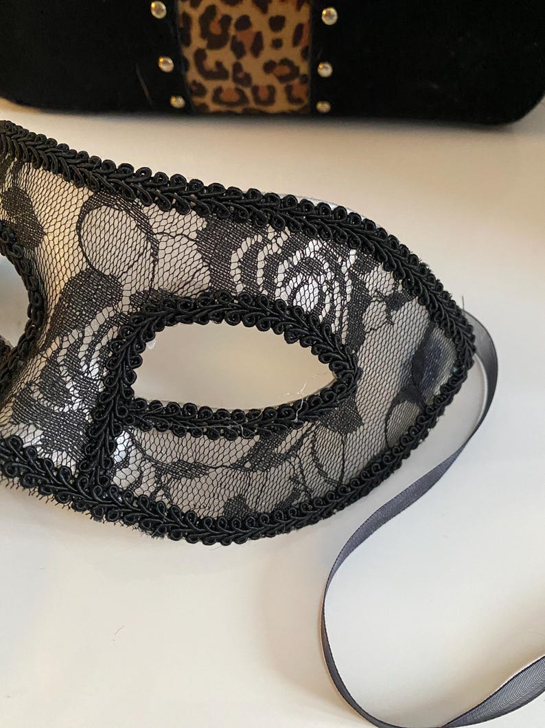 May include: A black lace masquerade mask with a black ribbon tie. The mask has a delicate floral pattern and a black satin trim.