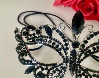 Puede incluir: Una máscara de carnaval negra con diseño de mariposa. La máscara está decorada con diamantes de imitación negros y tiene un marco de metal plateado. La máscara está unida a una banda elástica negra.