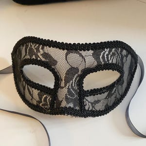 May include: A black lace masquerade mask with a black ribbon tie. The mask has a delicate floral pattern and is trimmed with a black cord.