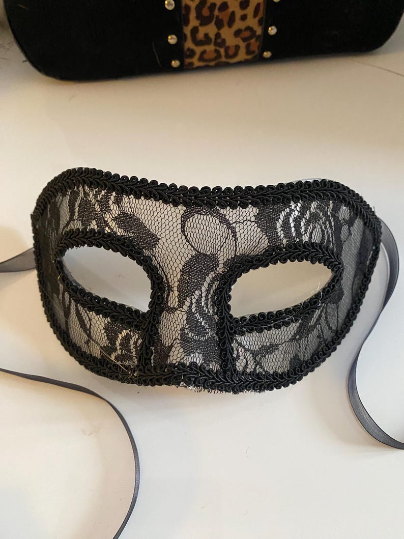 May include: A black lace masquerade mask with a black ribbon tie. The mask has a delicate floral pattern and is trimmed with a black cord.