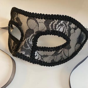 May include: A black lace masquerade mask with a black ribbon. The mask has a delicate floral pattern and a black trim.