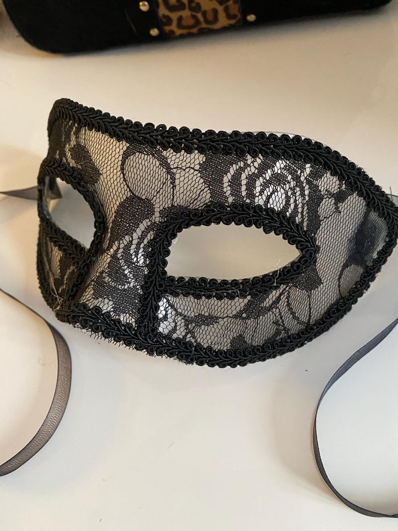 May include: A black lace masquerade mask with a black ribbon. The mask has a delicate floral pattern and a black trim.