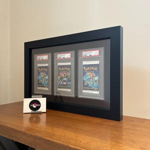 Op de afbeelding: Een zwart omkaderde display met drie Pokémon-ruilkaartpakketten. De pakketten zijn in beschermende houders geplaatst en voorzien van kleurrijke illustraties. Een kleine visitekaartje met een logo ligt op het houten oppervlak.