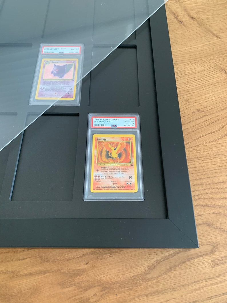 Puede incluir: Un marco negro con una parte superior de acr&iacute;lico transparente contiene una carta Pok&eacute;mon clasificada. La carta es un Moltres del set Fossil, clasificada NM-MT por PSA con una calificaci&oacute;n de 8. El n&uacute;mero de la carta es 113 y el n&uacute;mero de PSA es 54712276.