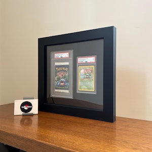 Puede incluir: Una exhibici&oacute;n enmarcada en negro con dos cartas de Pok&eacute;mon clasificadas. La carta de la izquierda es un paquete de refuerzo sellado con el logotipo de Pok&eacute;mon. La carta de la derecha es una carta de Bulbasaur clasificada. Una tarjeta de visita con un logotipo est&aacute; sobre la mesa.