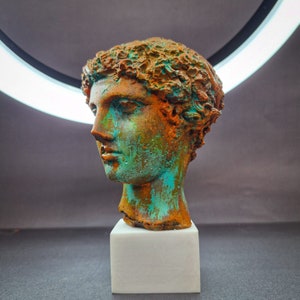 May include: A bronze bust of a person with a detailed hairstyle. The bust is painted with a green and brown patina. The bust is sitting on a white pedestal.