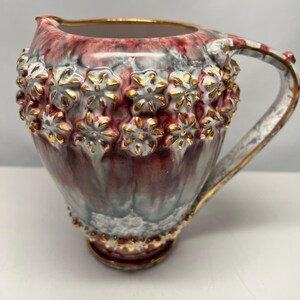 May include: A red and white ceramic pitcher with gold accents and a floral design. The pitcher has a handle and a rounded base.
