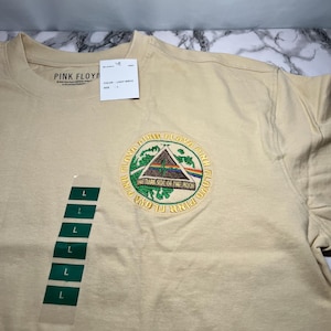 May include: A light birch-colored t-shirt featuring a circular embroidered patch with the text "PINK FLOYD" and "THE DARK SIDE OF THE MOON". The patch includes a triangle design with a rainbow and a mountain scene. The shirt has a size L tag.