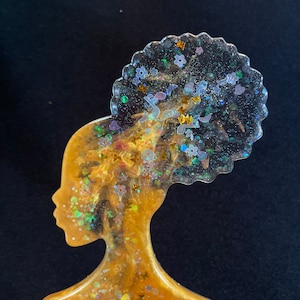 Op de afbeelding: Een gouden kam met een transparante hars bovenkant met een silhouet van een vrouw met een afro. De hars is gevuld met iriserende glitter en kleine bloemen.
