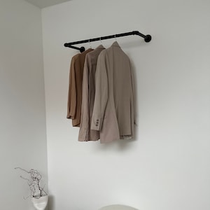Könnte beinhalten: Drei beige Jacken hängen an einer schwarzen Metallrohr-Kleiderstange, die an einer weißen Wand montiert ist. Die Kleiderstange befindet sich in einer Ecke des Raumes.