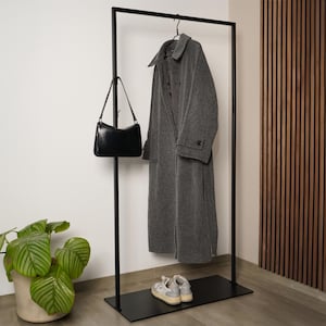 Op de afbeelding: Een zwarte metalen kapstok met een lange grijze jas aan een hanger. Een zwarte handtas hangt aan de kapstok en een paar grijze sneakers staat op de basis. Een plant in een pot is links zichtbaar.