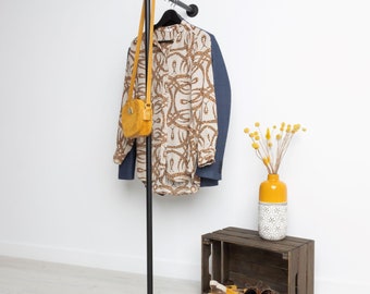 May include: A black metal clothing rack with a shirt, blazer, and purse. The shirt has a cream background with a brown pattern. A yellow purse hangs from the rack. A wooden crate holds shoes and a yellow vase with dried flowers.