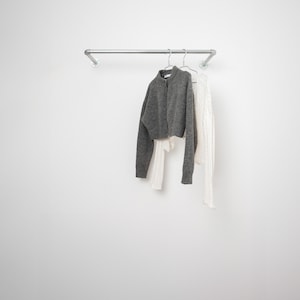 Peut inclure: Un cardigan gris et un pull blanc suspendus à un portant en tuyau d'argent sur un mur blanc.