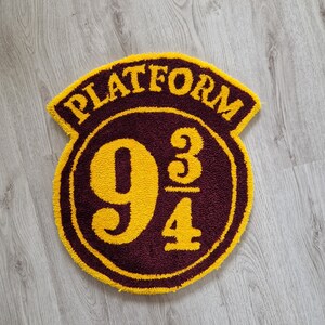 Peut inclure: Un tapis rond de couleur bordeaux avec une bordure jaune et les mots "Platform 9 3/4" en lettres jaunes.