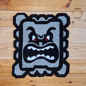 Op de afbeelding: Een grijs en zwart tapijt met een cartoon gezicht met een boze uitdrukking. Het gezicht heeft rode ogen en een witte mond met tanden.