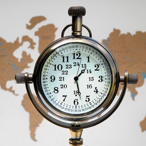 Puede incluir: Un reloj de lat&oacute;n de estilo vintage con esfera blanca y n&uacute;meros negros. El reloj tiene un dise&ntilde;o redondo con asa y soporte. La esfera muestra formatos de hora de 12 y 24 horas. El fondo es un mapa del mundo. El reloj mide unos 15 cm de di&aacute;metro.