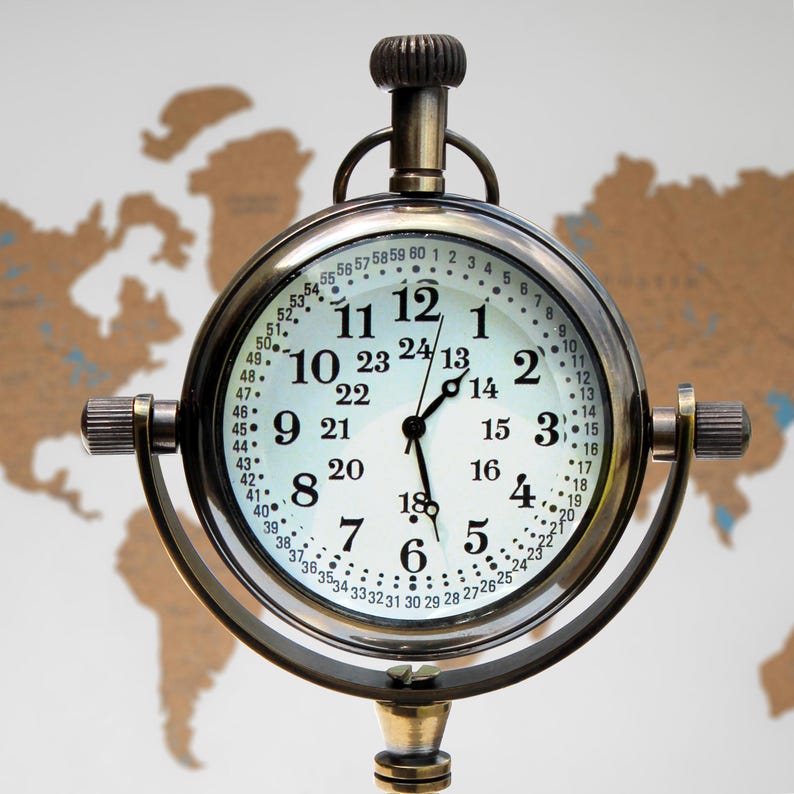 Puede incluir: Un reloj de lat&oacute;n de estilo vintage con esfera blanca y n&uacute;meros negros. El reloj tiene un dise&ntilde;o redondo con asa y soporte. La esfera muestra formatos de hora de 12 y 24 horas. El fondo es un mapa del mundo. El reloj mide unos 15 cm de di&aacute;metro.