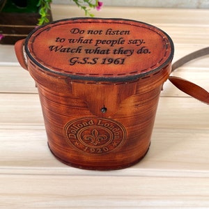 K&ouml;nnte beinhalten: Eine braune, zylindrische Lederbox mit einem eingravierten Zitat auf der Oberseite: "H&ouml;re nicht auf das, was die Leute sagen. Beobachte, was sie tun. G.S.S 1961". Die Box hat ein kreisf&ouml;rmiges Logo an der Seite, auf dem "Dollond London 1920" steht.