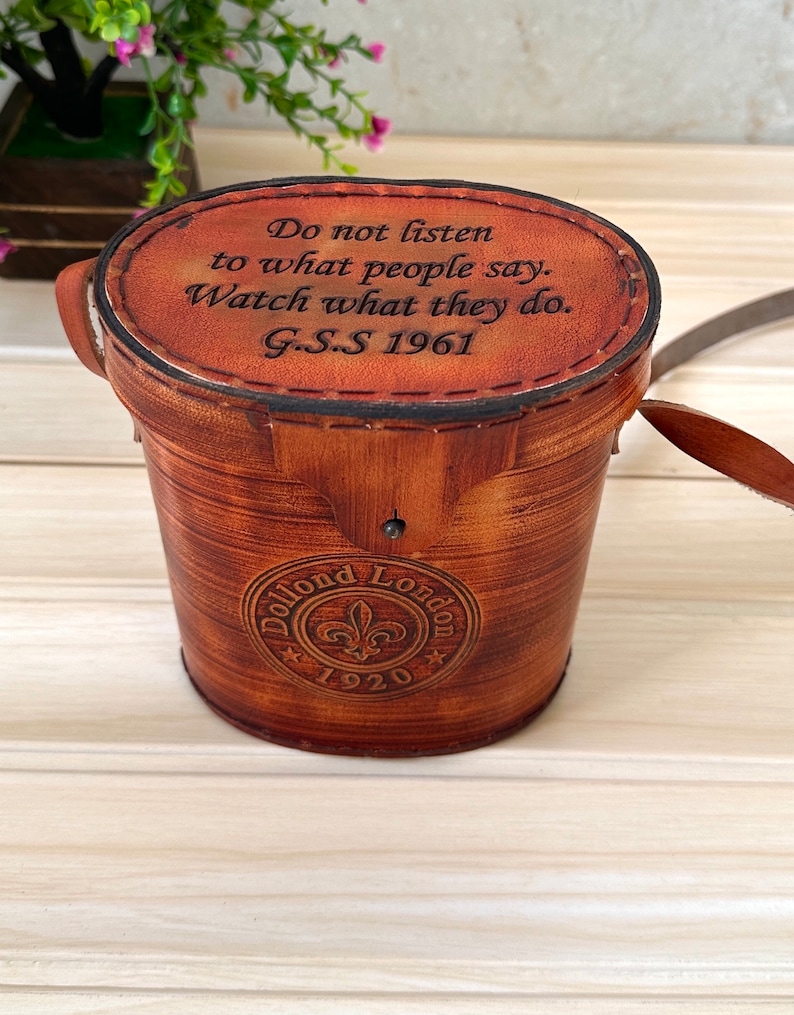 K&ouml;nnte beinhalten: Eine braune, zylindrische Lederbox mit einem eingravierten Zitat auf der Oberseite: "H&ouml;re nicht auf das, was die Leute sagen. Beobachte, was sie tun. G.S.S 1961". Die Box hat ein kreisf&ouml;rmiges Logo an der Seite, auf dem "Dollond London 1920" steht.