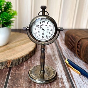 Puede incluir: Un reloj de escritorio de lat&oacute;n de estilo antiguo con una esfera blanca y n&uacute;meros negros. El reloj tiene una esfera redonda con un bisel detallado y un soporte con una base ornamentada. El reloj mide aproximadamente 15 cm de alto.