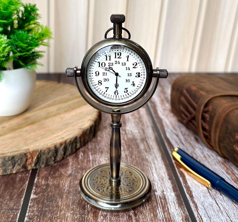 Puede incluir: Un reloj de escritorio de lat&oacute;n de estilo antiguo con una esfera blanca y n&uacute;meros negros. El reloj tiene una esfera redonda con un bisel detallado y un soporte con una base ornamentada. El reloj mide aproximadamente 15 cm de alto.
