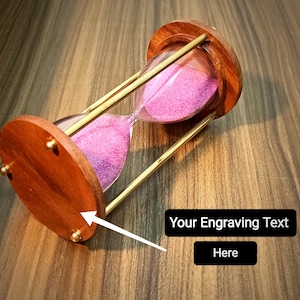Könnte beinhalten: Eine Sanduhr aus Holz mit einem Messingrahmen und rosa Sand. Die Sanduhr steht auf einer Holzoberfläche.  "Your Engraving Text Here" ist auf einem schwarzen Rechteck gedruckt.