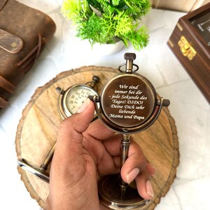 Puede incluir: Una br&uacute;jula de lat&oacute;n vintage con un soporte, con una esfera circular y texto grabado. La br&uacute;jula se sostiene en una mano, con una rodaja de madera y un peque&ntilde;o reloj en el fondo. El texto en la br&uacute;jula dice "Wir sind immer bei dir - jede Sekunde des Tages! DIDO! Deine Dich sehr liebende Mama und Papa."