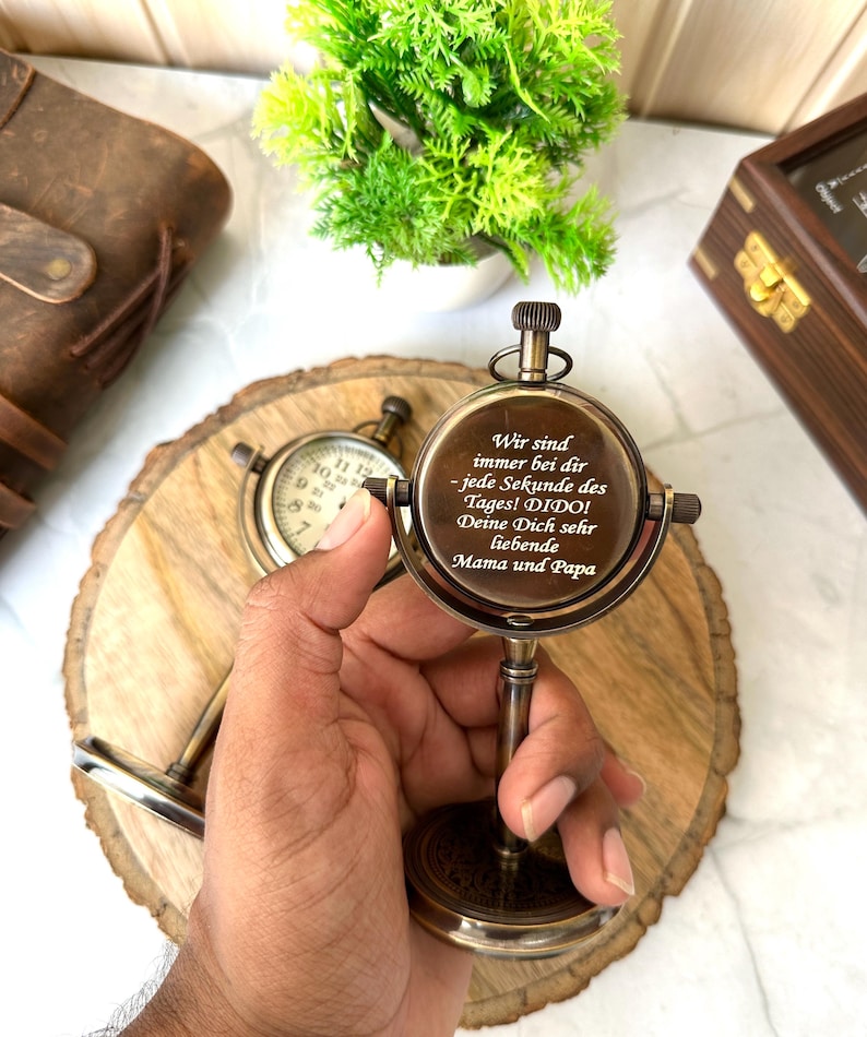 Puede incluir: Una br&uacute;jula de lat&oacute;n vintage con un soporte, con una esfera circular y texto grabado. La br&uacute;jula se sostiene en una mano, con una rodaja de madera y un peque&ntilde;o reloj en el fondo. El texto en la br&uacute;jula dice "Wir sind immer bei dir - jede Sekunde des Tages! DIDO! Deine Dich sehr liebende Mama und Papa."